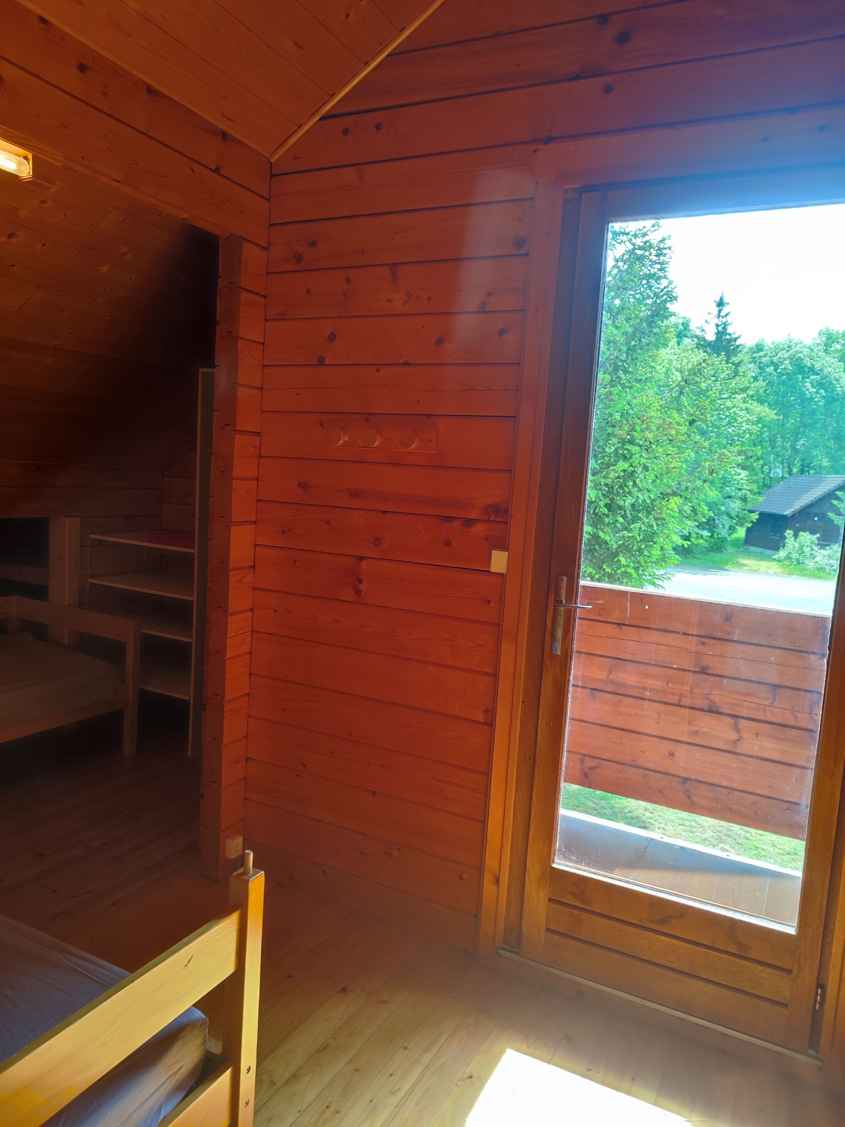 A wooden cabin room with a bunk bed, a small table, and a window with a view of a forest and a cabin.