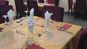 Un espace repas avec des tables dressées pour quatre personnes, chacune étant équipée de couverts, de serviettes et de verres. Des chaises sont disposées autour des tables, et la pièce est dotée d’une grande fenêtre avec des stores.