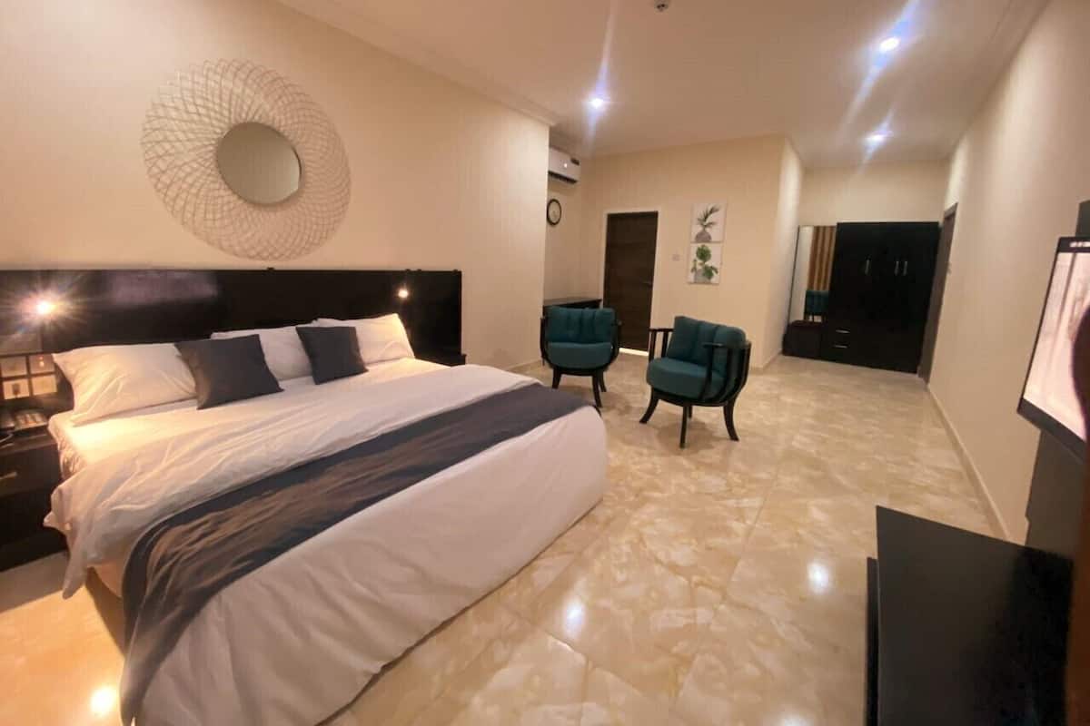 A modern hotel room with a large bed, two armchairs, a round mirror, and a flat-screen TV.