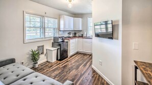 Un espacio habitable compacto con kitchenette, un sofá gris con capitonado, una planta en maceta y un televisor de pantalla plana montado en la pared.