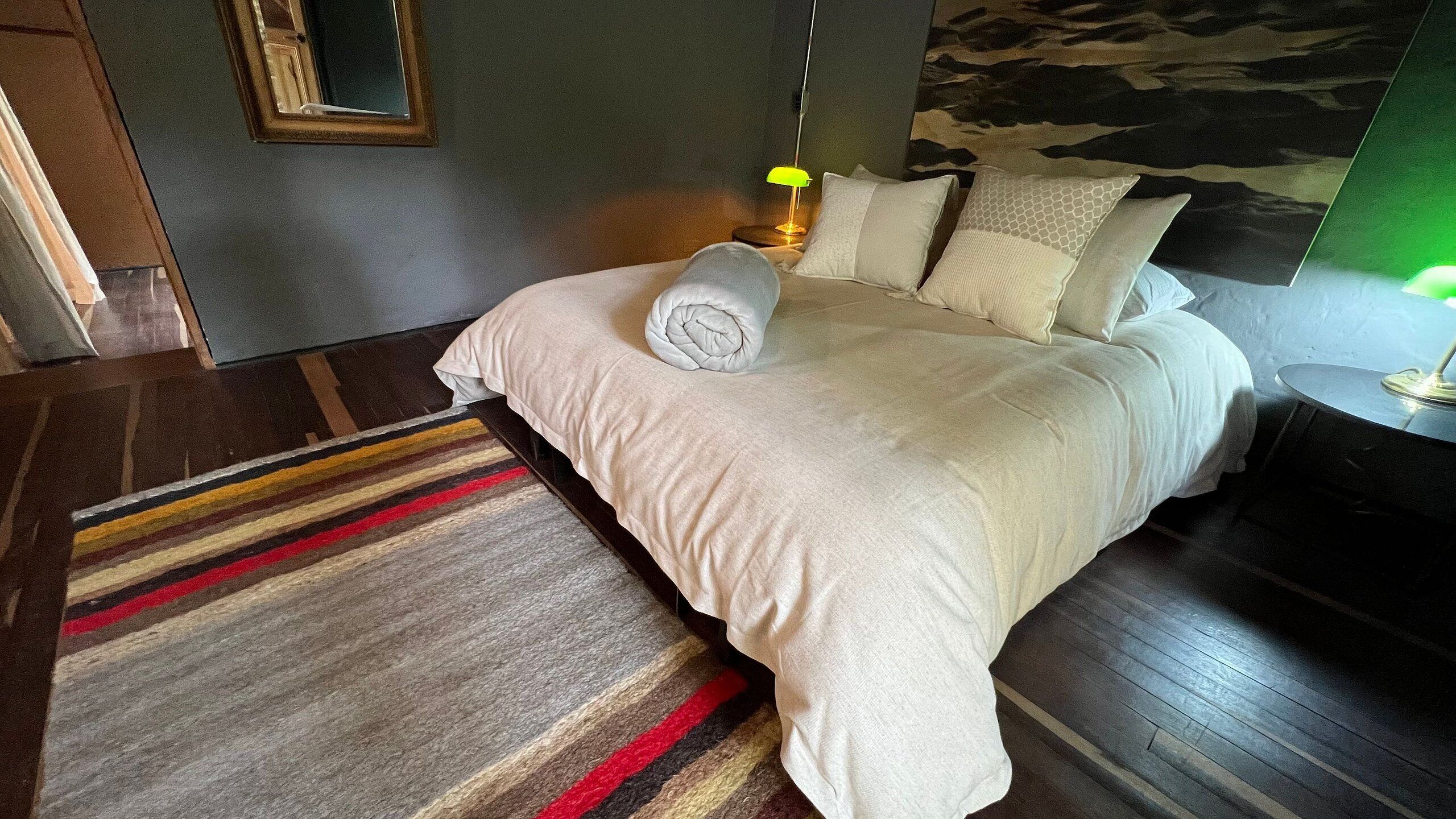 A neatly made bed with a rolled towel, a striped rug, and a nightstand with a lamp.