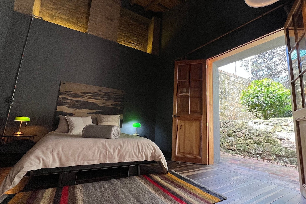 A bedroom with a bed, bedside lamps, a headboard with a painting, a wooden door, and a view of a garden.
