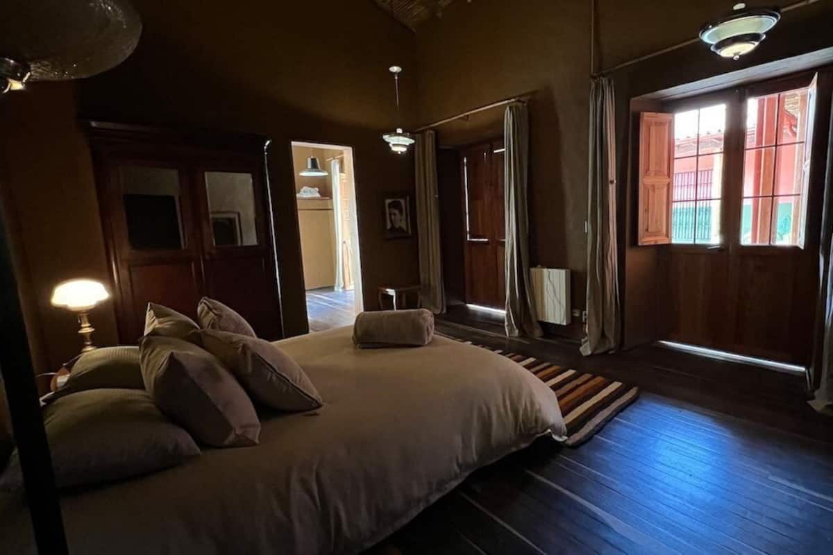 A bedroom with a large bed, wooden flooring, and a view of the outside through a window.