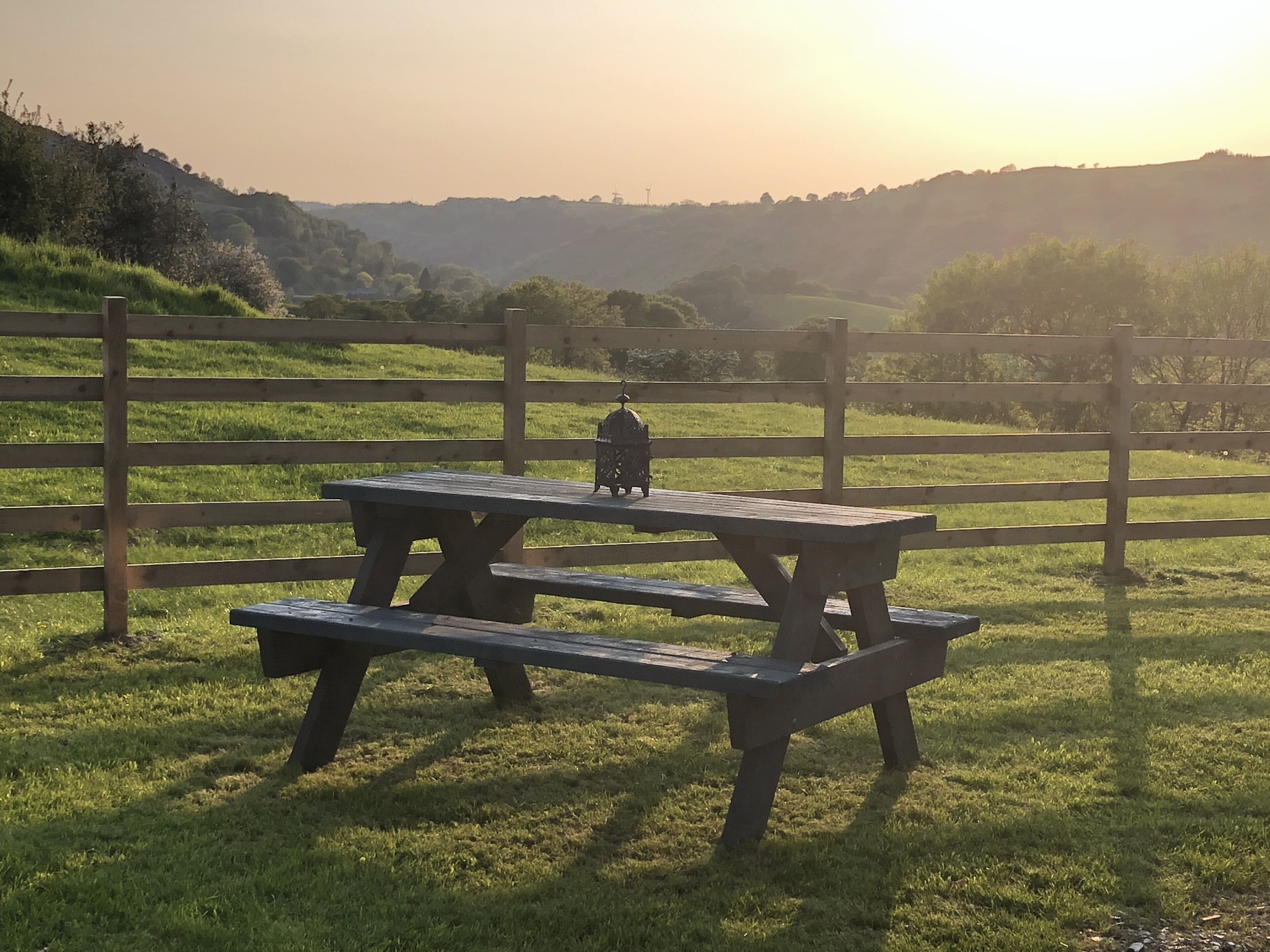 A picnic table with a lantern in a grassy field, surrounded by a wooden fence.