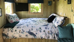 A wooden cabin bedroom with a bed, two windows, a TV, and a small table.