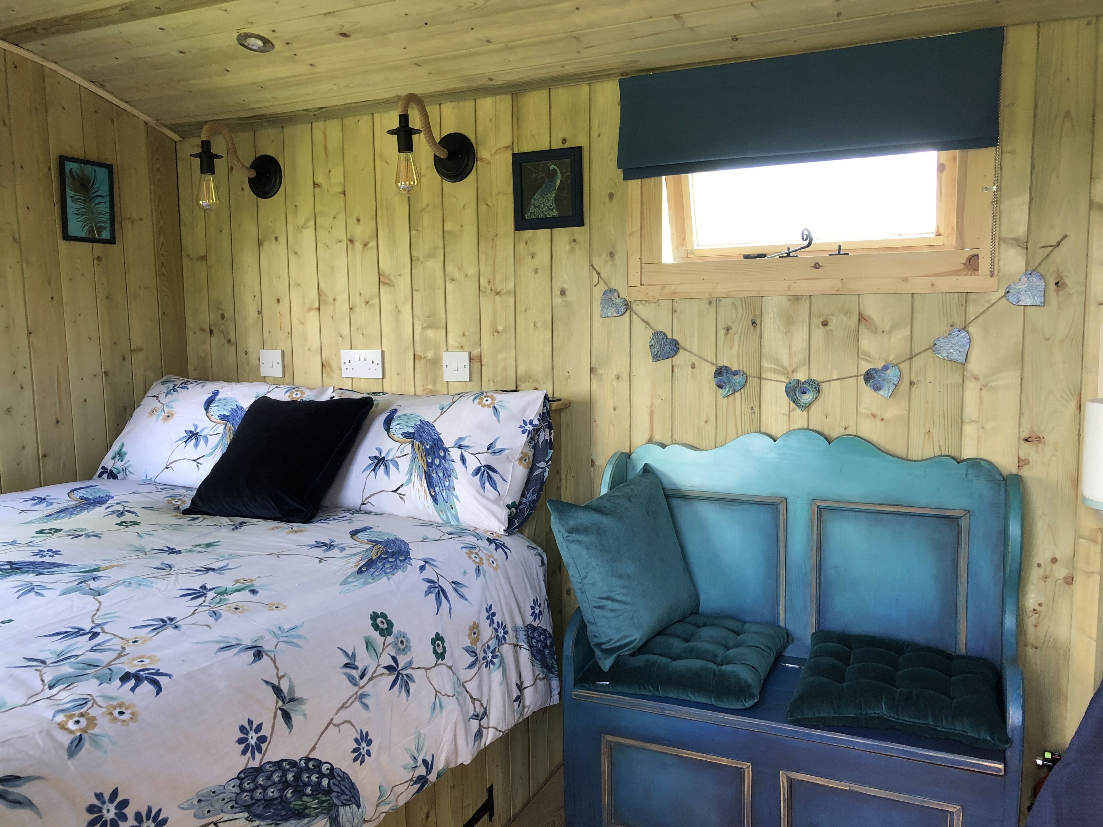 A wooden room with a bed and a daybed. There are wall-mounted light fixtures and a window with a curtain.