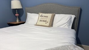 Ein ordentlich bezogenes Bett mit weißen Leinentüchern, einer flauschigen grauen Decke und einem Kopfkissen mit der Aufschrift 'Zuhause ist da, wo das Herz ist'.