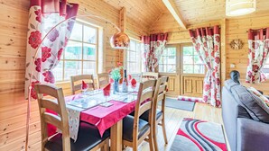 L’intérieur d’un chalet en bois comprend une table à manger pour deux personnes, des chaises en bois, un canapé et une grande fenêtre avec des rideaux.