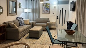 Una sala de estar con sofá, reposapiés, mesa de vidrio y sillas. Hay un ventilador de techo, un cuadro y un televisor en la pared.