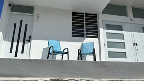 Un edificio de fachada blanca, dos sillas azules en el porche, una puerta de garaje con el número 366, y una ventana con persianas.