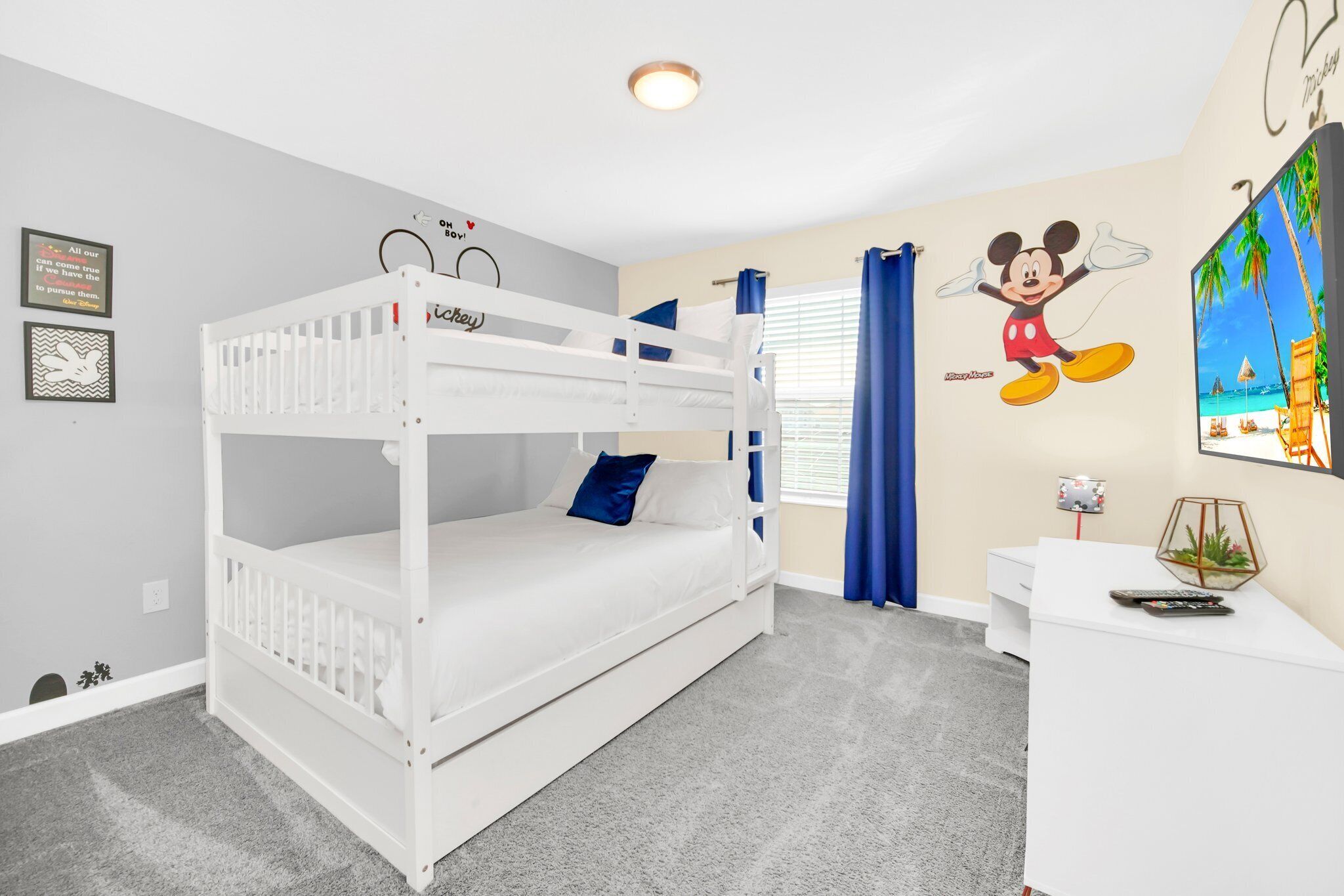 Een slaapkamer met stapelbed, een wit kleurenschema, een Mickey Mouse muurstickers, een klein bureau met een plant en een televisie rechts.