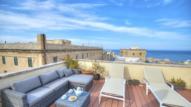Une terrasse sur le toit, avec un canapé en osier, deux fauteuils, une table avec une théière et des assiettes, et des plantes en pot.