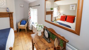 A bedroom with a large mirror, a bed with blue bedding, a wooden dresser, and a wicker chair. - The Cottage (Stratford-upon-Avon)