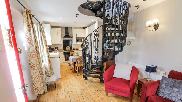 A modern living room with a red sofa, a wooden floor, a spiral staircase, and a kitchen in the background. - The Cottage (Stratford-upon-Avon)