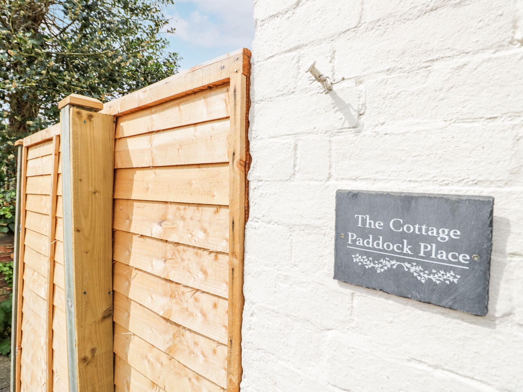 A wooden gate and a sign reading "The Cottage Paddock Place" on a white wall.