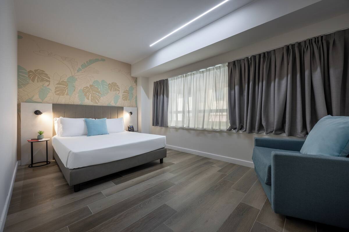 A modern bedroom with a large bed, a bedside table, a chair, a window with curtains, and a wall mural.