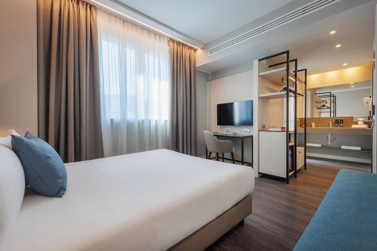A hotel room with a bed, a desk, a chair, a TV, and a bathroom with a mirror and shelves.