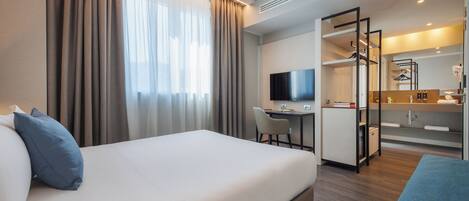 A hotel room with a bed, a desk, a chair, a TV, and a bathroom with a mirror and shelves.
