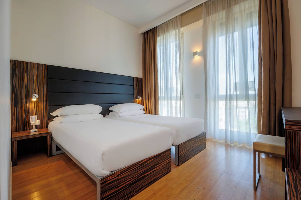 A hotel room with two beds, wooden flooring, a dark headboard, and a view of the outdoors through sheer curtains.