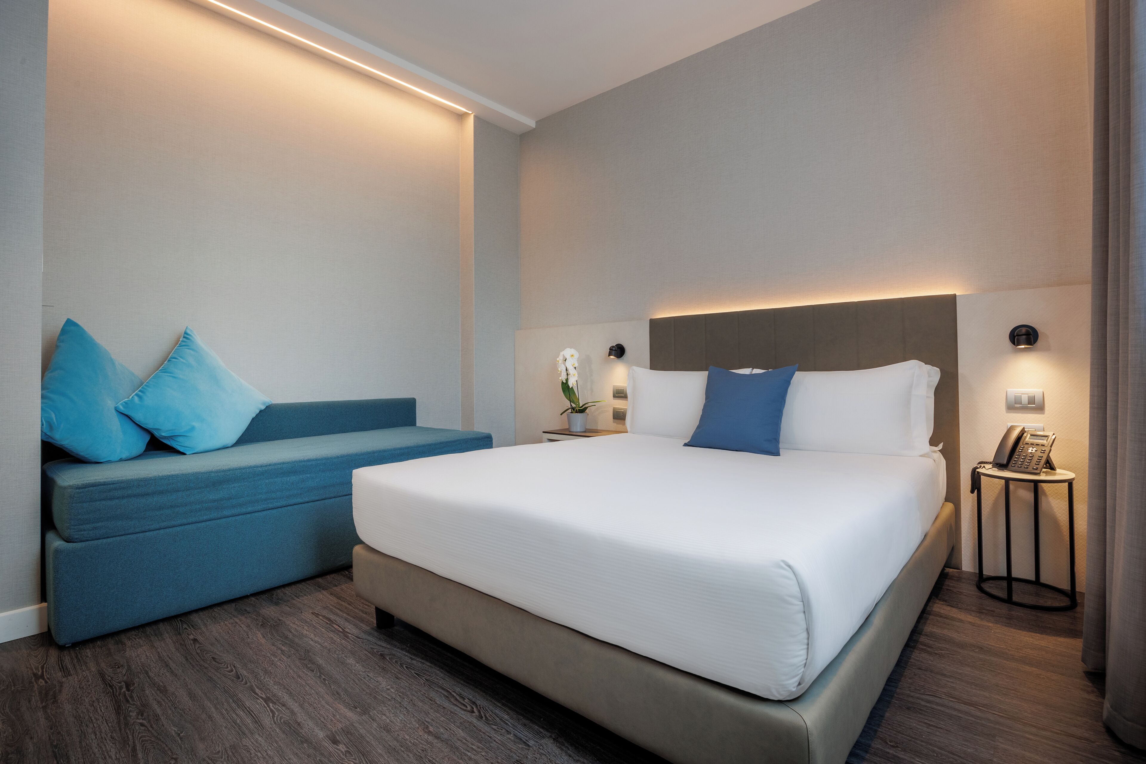 Una habitación de hotel moderna con una cama grande, un sofá azul y una mesita de noche con un teléfono.