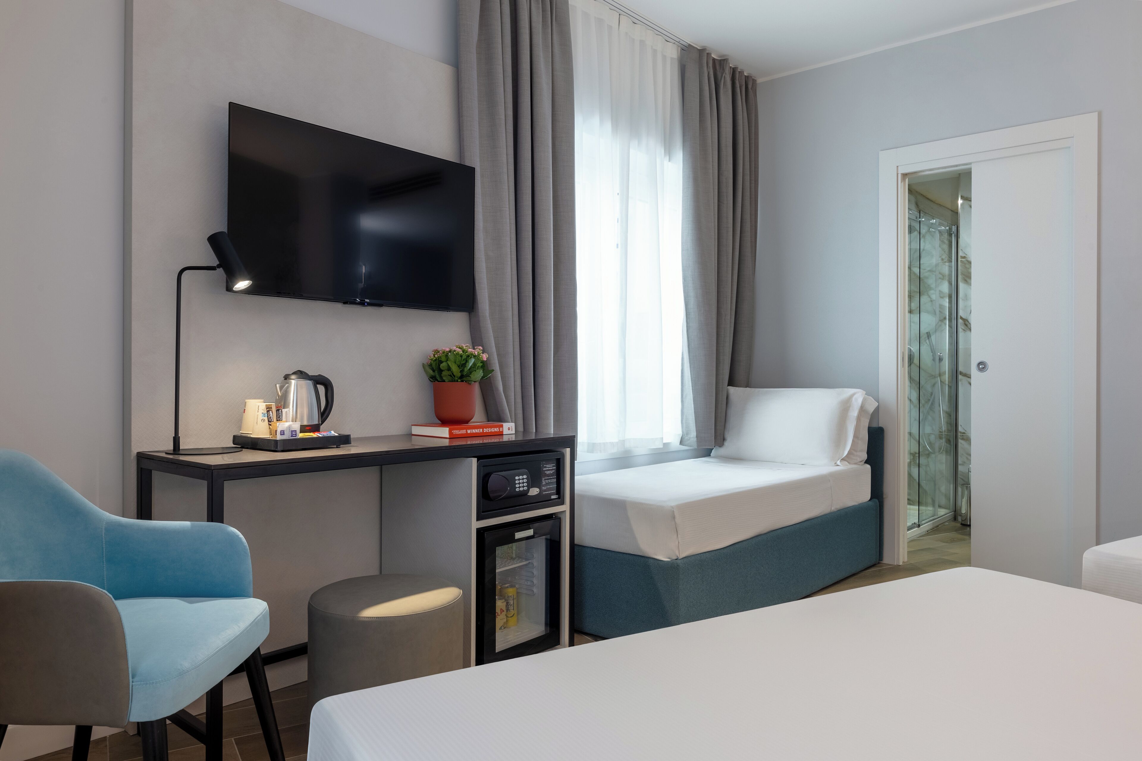 Ein modernes Hotelzimmer mit flachbildfernseher an der Wand, einem Schreibtisch mit Kaffeemaschine, einem kleinen Kühlschrank, einem Bett mit blauer Kopfteil und einer Glastür zum Balkon.