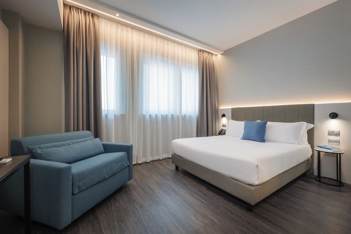 A hotel room with a bed, a blue sofa, a bedside table, and a window with curtains.