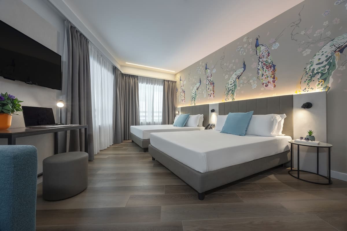 A hotel room with two beds, a desk with a laptop, a TV, and a wall with peacock murals.