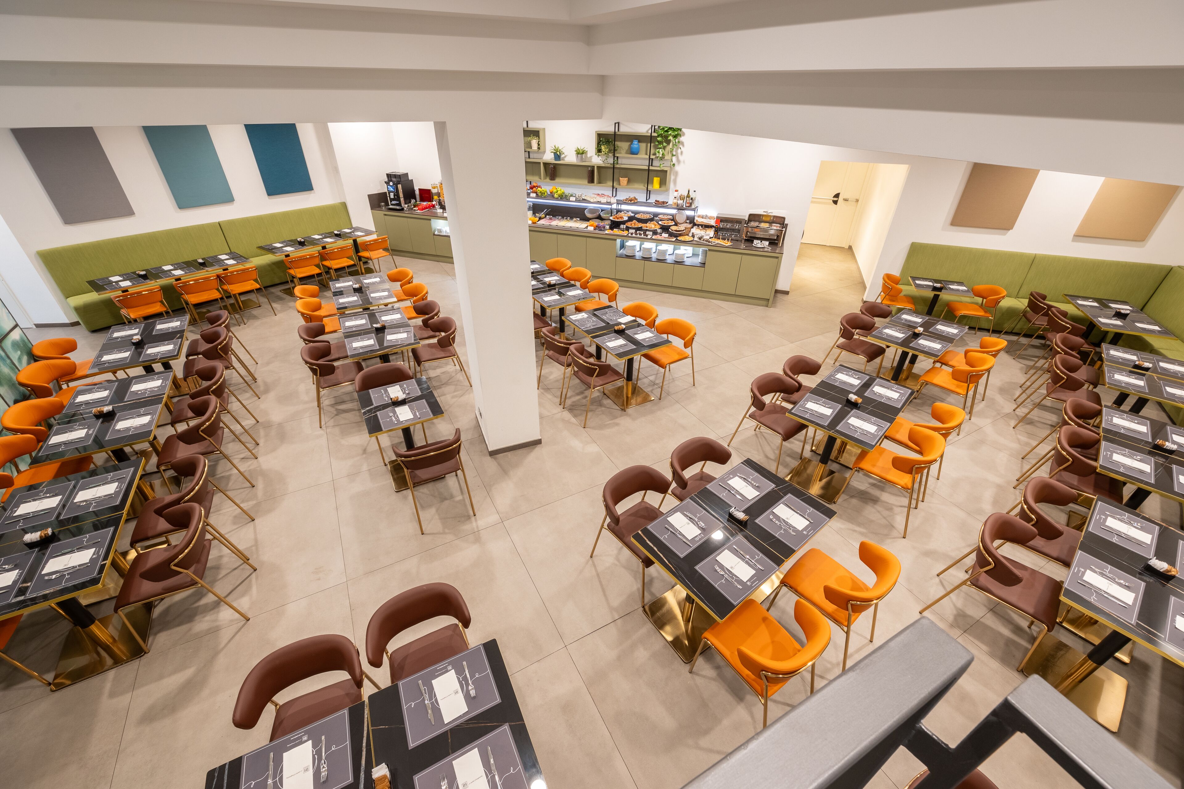 A modern dining area with multiple tables and chairs, a central counter with food display, and green cushioned seating areas.