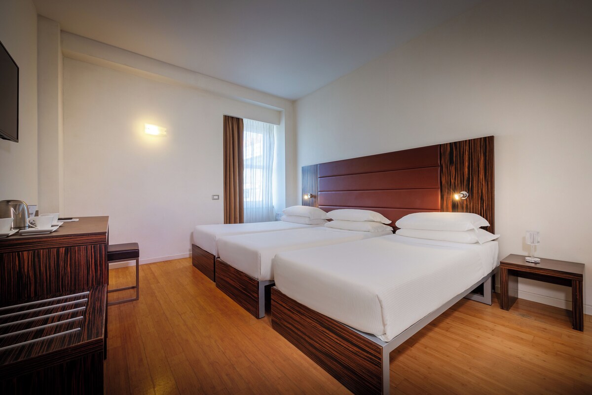 A hotel room with two beds, a wooden dresser, a desk with a chair, and a nightstand with a lamp.