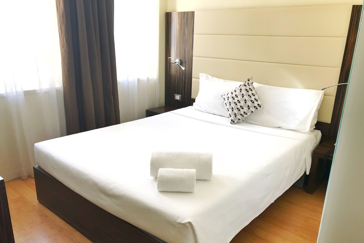 A neatly made bed with white linens and a patterned pillow, a bedside table, and a wooden floor.