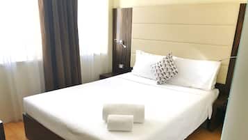 Cama bem arrumada com lençóis brancos e almofada estampada, mesa de cabeceira e piso de madeira.