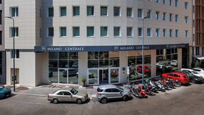 A modern hotel building with a curved facade, multiple windows, and a parking area with cars and motorcycles. - IH Hotels Milano Centrale (Milan)