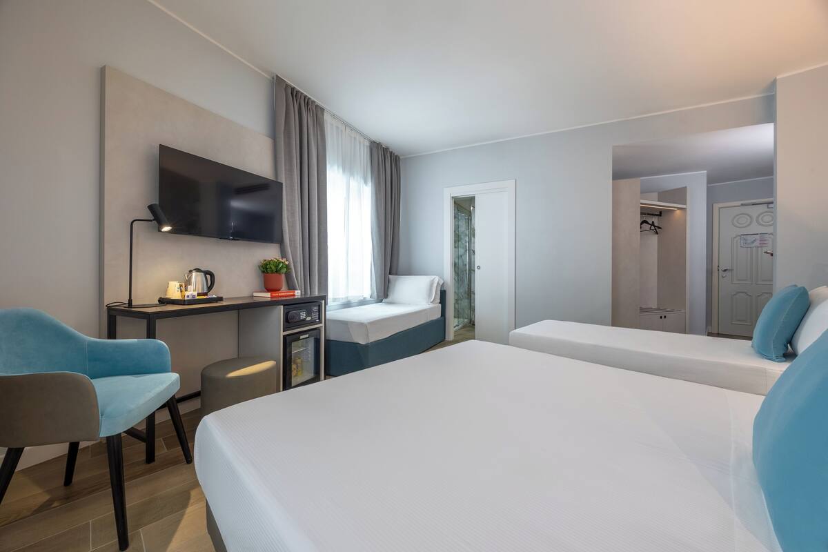 A hotel room with a bed, a desk with a TV, a chair, a small table with a plant, and a door leading to another room.