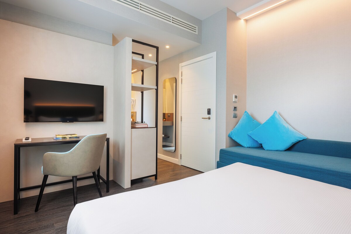 A hotel room with a bed, a desk, a chair, a TV, a sofa, and a door leading to a bathroom.