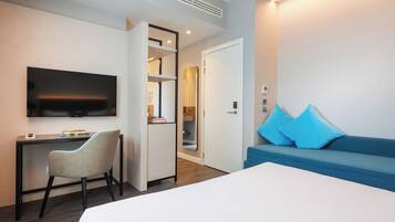 A hotel room with a bed, a desk, a chair, a TV, a sofa, and a door leading to a bathroom.