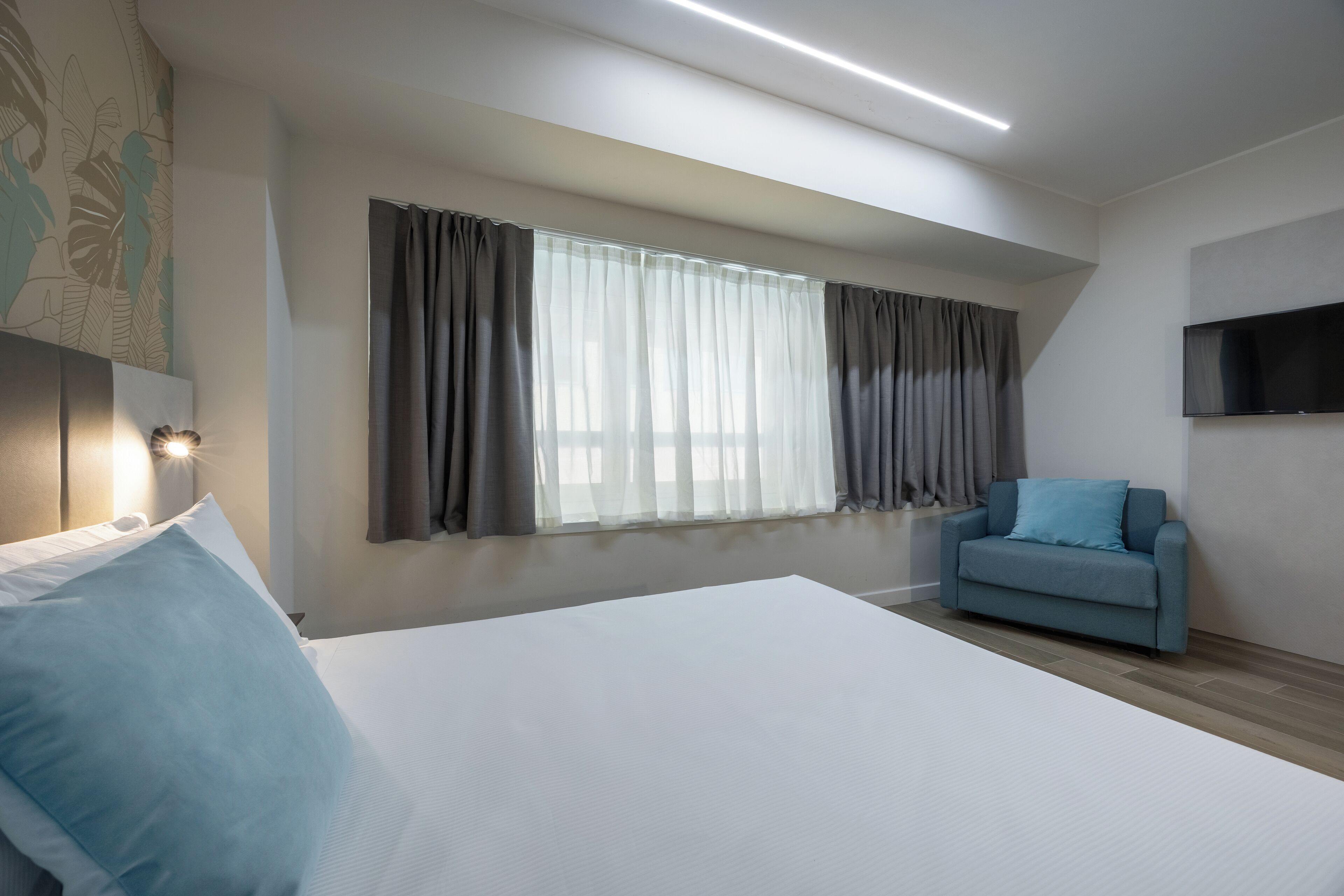 Una camera d'albergo con un letto grande, una poltrona blu, una televisione a schermo piatto e una finestra con tende.