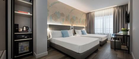 Una habitación de hotel moderna con dos camas, un minibar, un escritorio con silla y un amplio ventanal con cortinas.