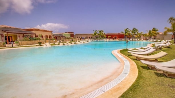Piscina com espreguiçadeiras, palmeiras e edifícios com telhados de terracota.