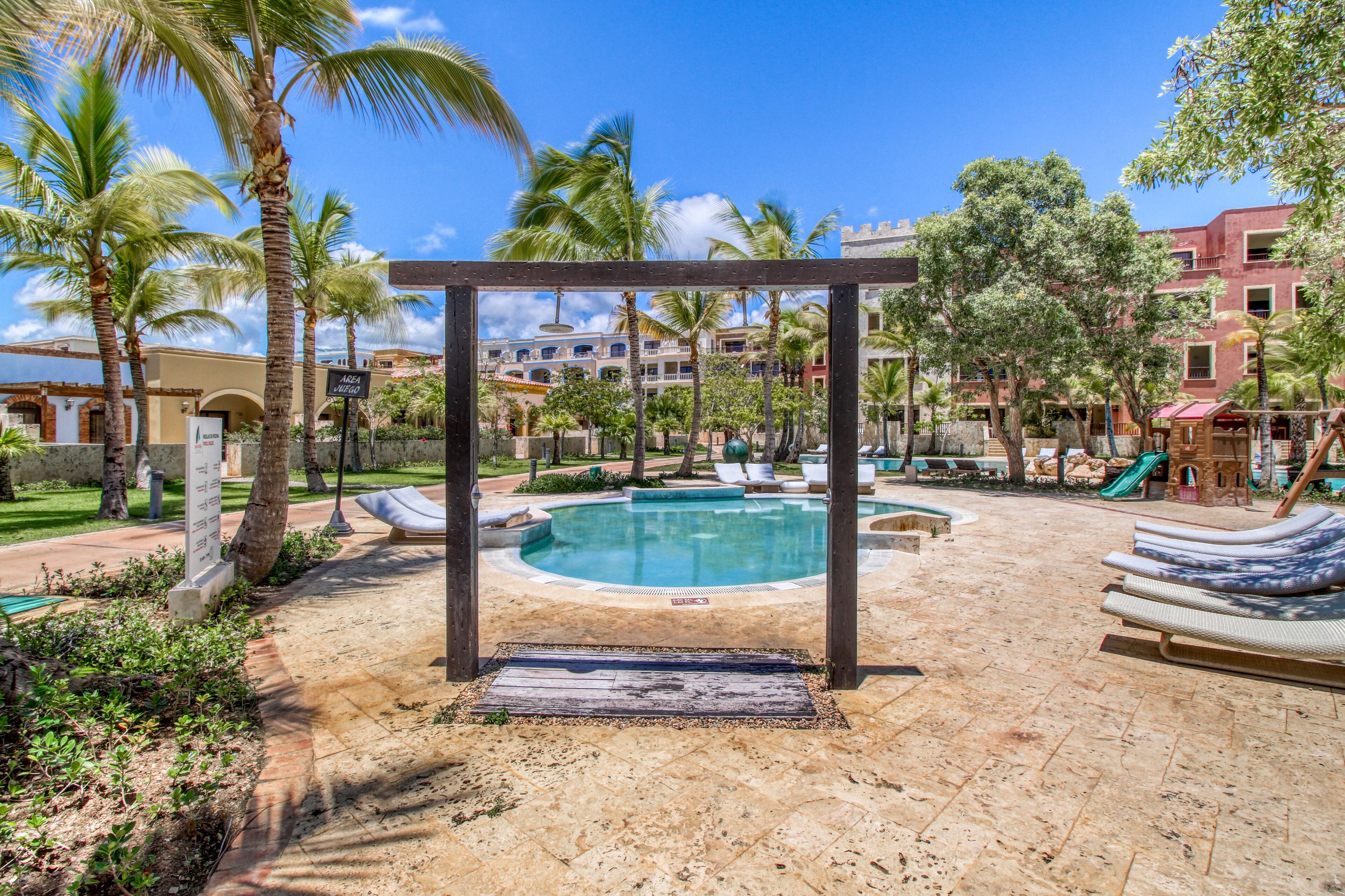 Un área de piscina con sillones de descanso, palmeras y una estructura de pérgola.