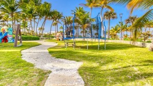Un sendero conduce a un parque infantil con un tobogán y estructuras para escalar, rodeado de palmeras y un cielo despejado.