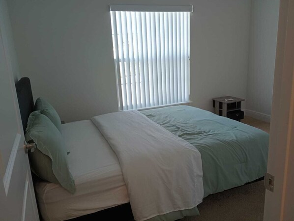 A bedroom with a bed, a window with blinds, a nightstand, and a small table.