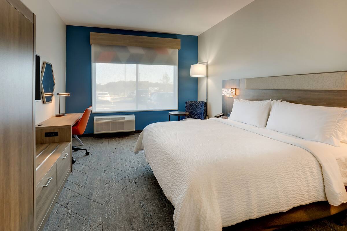 A hotel room with a large bed, a desk, a chair, a window with a view of the parking lot, and a wall-mounted TV.