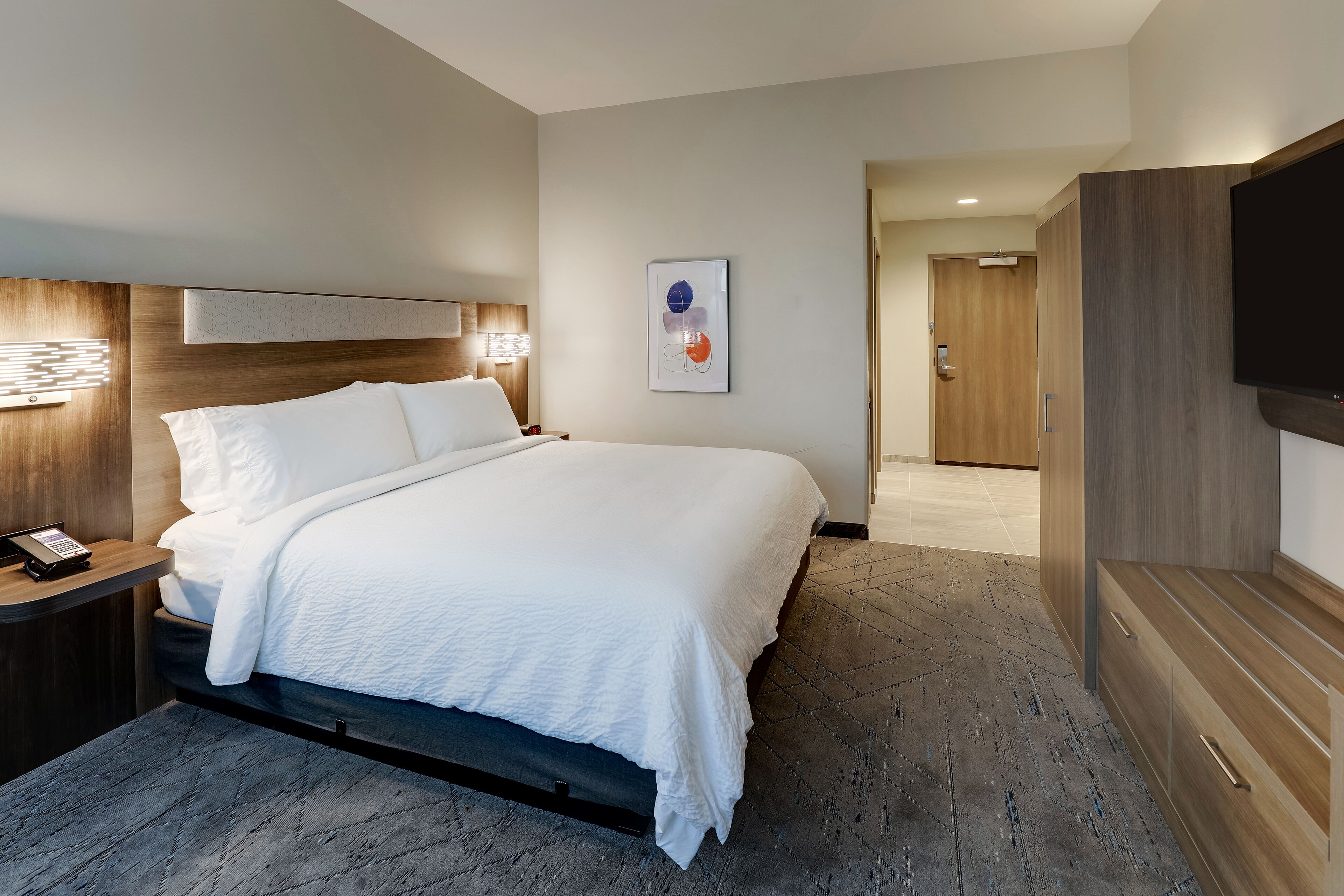A modern hotel room with a large bed, bedside tables, a flat-screen TV, and a framed artwork on the wall.