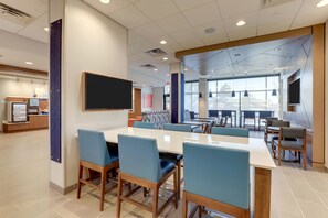 A modern dining area with a long table, blue chairs, and a flat-screen TV mounted on the wall. - Holiday Inn Express Holly Springs Raleigh Area by IHG (Holly Springs)
