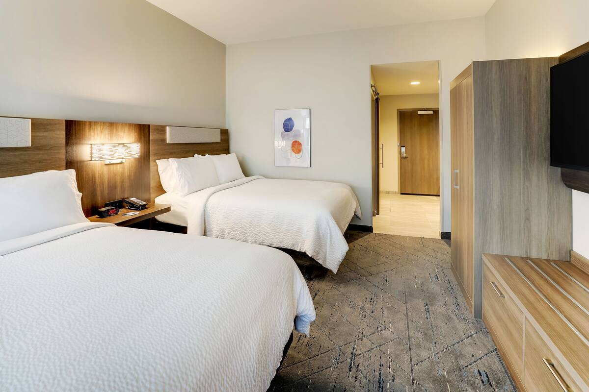 A hotel room with two beds, a wooden headboard, a flat-screen TV, a bedside table with a lamp, and a framed artwork on the wall.