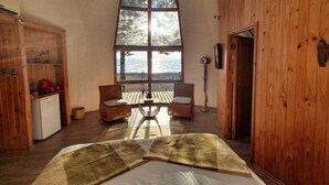 A room with a large window, wooden walls, a bed, two chairs, a small table, and a refrigerator. - Magma Lodge Pucon (Pucón)