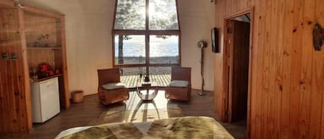 A room with a large window, wooden walls, a bed, two chairs, a small table, and a refrigerator.