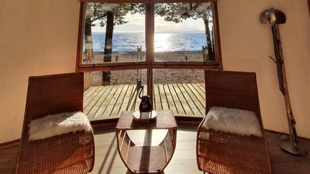 A room with a large window overlooking a beach, two wicker chairs, a small table, and a lamp. - Magma Lodge Pucon (Pucón)