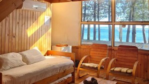A cozy cabin interior with a wooden bed, a small table, and a window overlooking a forest. - Magma Lodge Pucon (Pucón)