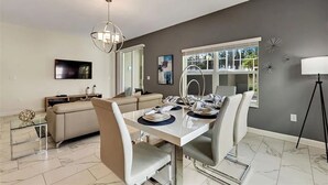 A modern dining area with a white table set for four, a sofa, and a TV mounted on the wall. - CG236OCA - Luxury 5BR-4BA Villa with Private Pool (Davenport)